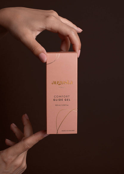 Hand holding a pink box of Augusta Comfort Glide Gel against a dark background